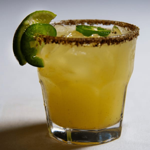A vibrant yellow cocktail with ice, garnished with lime and jalapeño slices, rimmed with spices.
