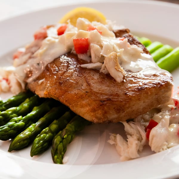 Pan-seared fish topped with crab meat, diced tomatoes, and a lemon wedge, served with asparagus.