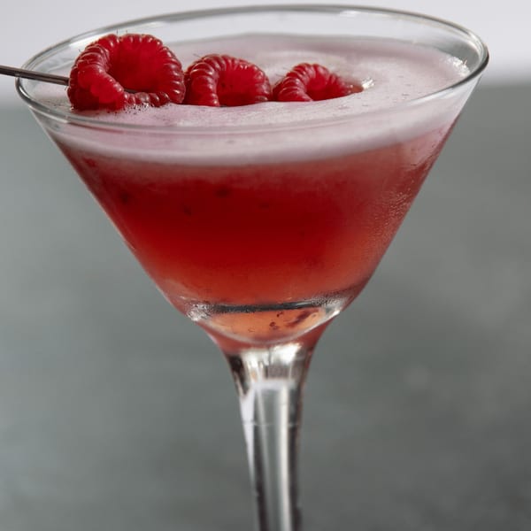 A vibrant red cocktail in a martini glass, garnished with three fresh raspberries on a skewer.