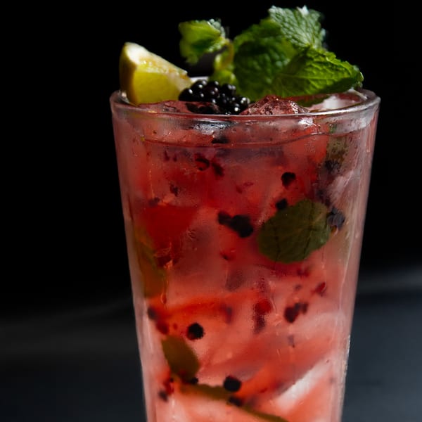 A vibrant red cocktail with blackberries, mint, and a lime wedge in a tall glass.