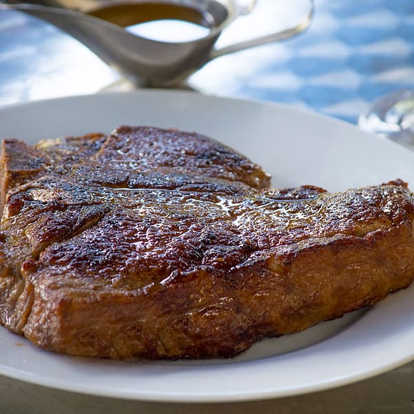 A perfectly seared steak, glistening with juices, rests on a white plate with a gravy boat in the background.