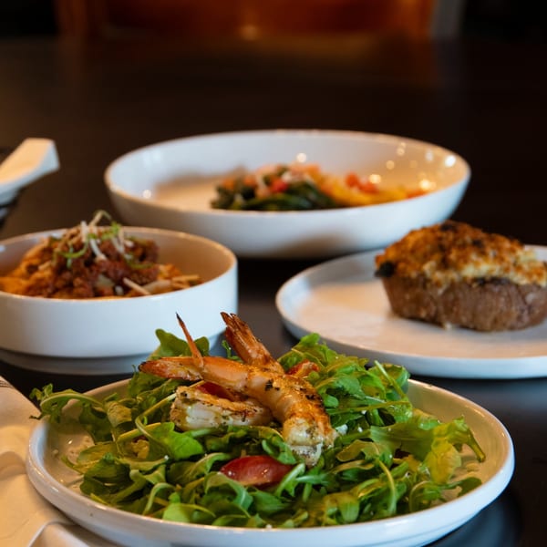 Grilled shrimp over a bed of arugula, with pasta and a stuffed mushroom in the background.